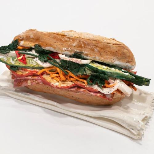 Image of ceramic Bánh mì sculpture on a white background. Some ingredients are identifiable as shredded carrot, chilli, coriander, salami, onion, and cucumber.  