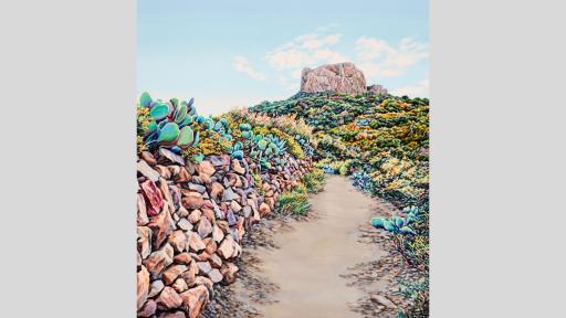 A dirt path leads uphill toward a large rock. A mixture of seaside and desert plants line the path on either side. 