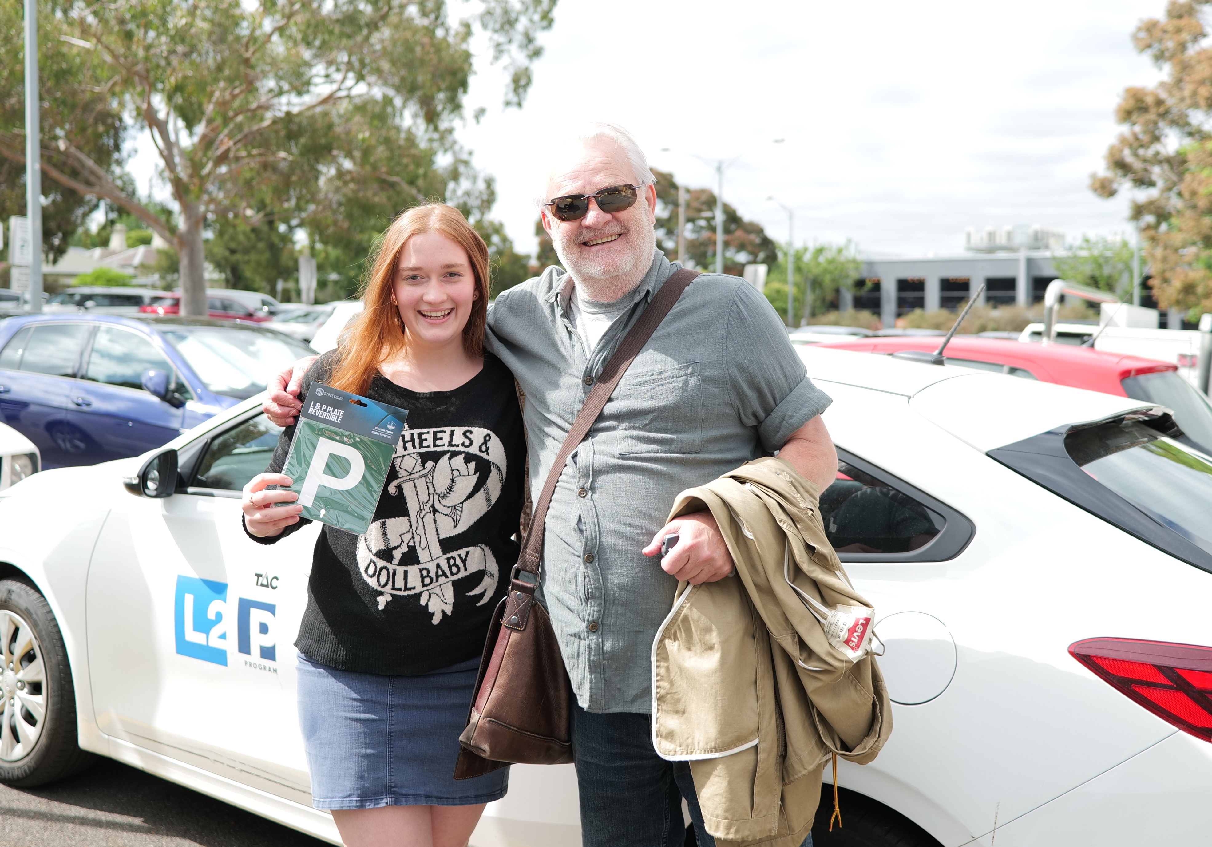 Young woman holding a P plate next to older man and in front of a car