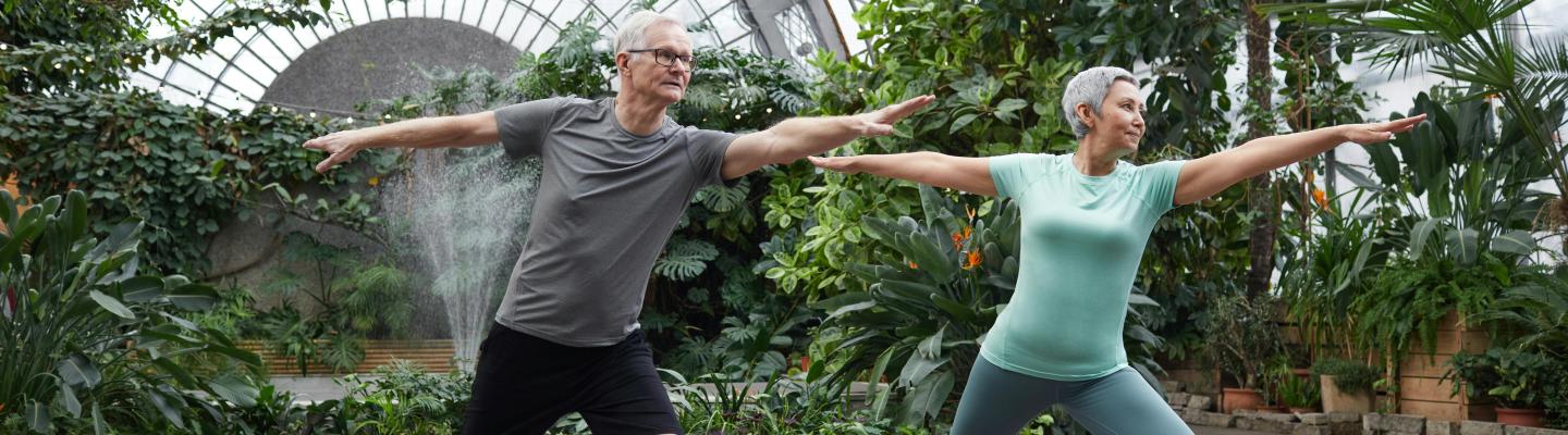 Two older adults doing standing forward stretch with knees apart, arms straight. They are standing on fitness mats in front of an abundance of green plants.