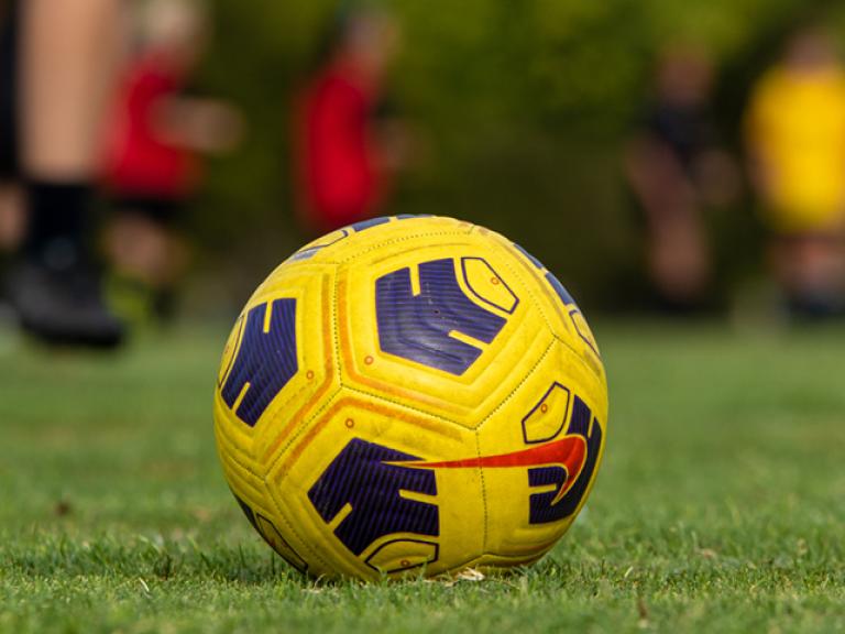 A black and yellow soccer ball sits on grass with several players in the background.