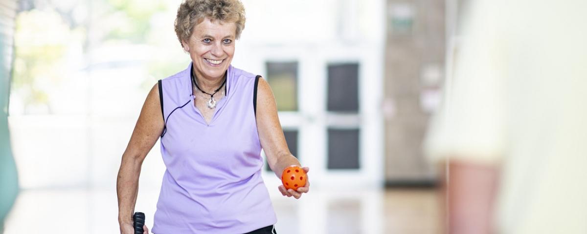 An older woman wearing a purple singlet holding a plastic ball with holes in it.