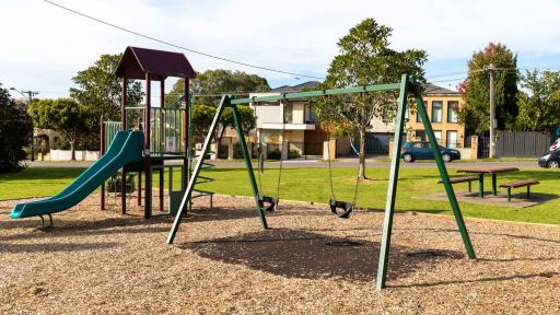 Playground with swings and small climbing feature, next to a table and chair set. There is a road, small trees and houses in the distance.