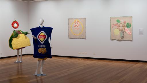 Two mannequins wearing colorful outfits stand in front of a gallery wall with three colorful artworks.