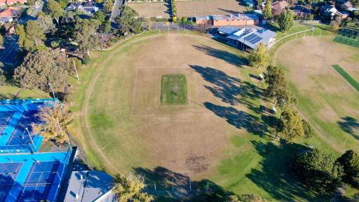 An aerial view of a sports oval. AFL posts and line markings are visible. There are blue tennis courts on the left, and another sports oval to the right. 