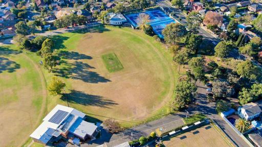 An aerial view of a sports oval. It is surrounded by trees and is next to a blue tennis courts. 