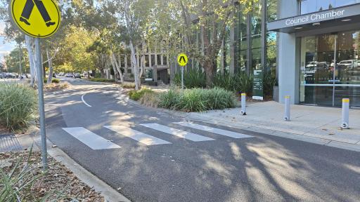 a pedestrian (zebra) crossing on a sun-dappled street leading to a building with a sign saying Council Chamber