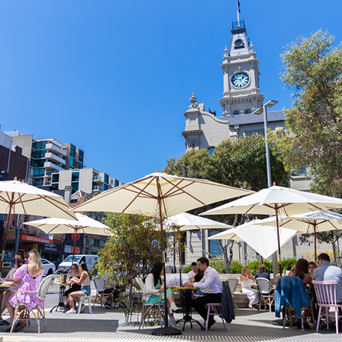 Outdoor tables and chairs sheltered by large umbrellas on a sunny day. In the background the clocktower of Hawthorn Arts Centre can be seen.