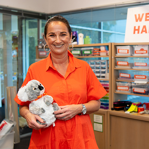 Amanda Mansie stands in an orange top and with a koala teddy smilling at the camera
