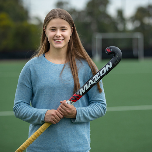 a girl wearing a blue jumper stands on a hockey pitch holding her hockey stick