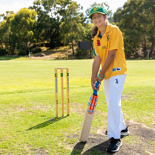 Young cricketer holding a cricket bat and standing in front of stumps.
