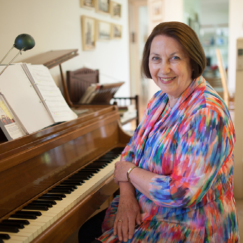 A woman wearing a colourful top sits in front of a piano and smiles at the camera