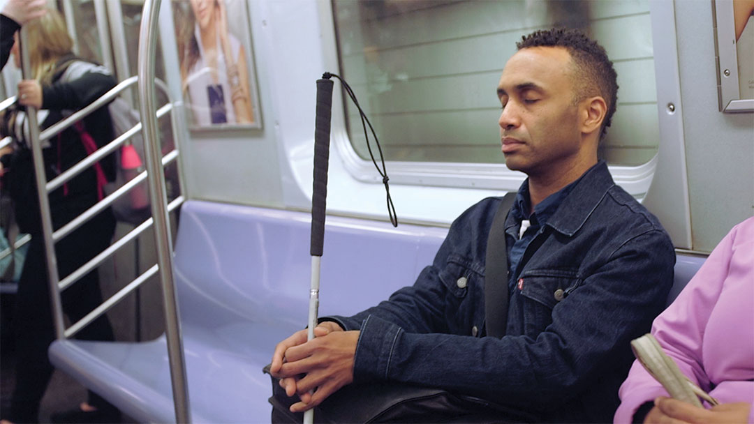 A Black man sits with his eyes closed on public transport while holding a cane for vision-impaired people