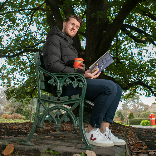 A person in puffer jacket with orange keep cup sitting on a park bench reading a book with a large tree filling the background