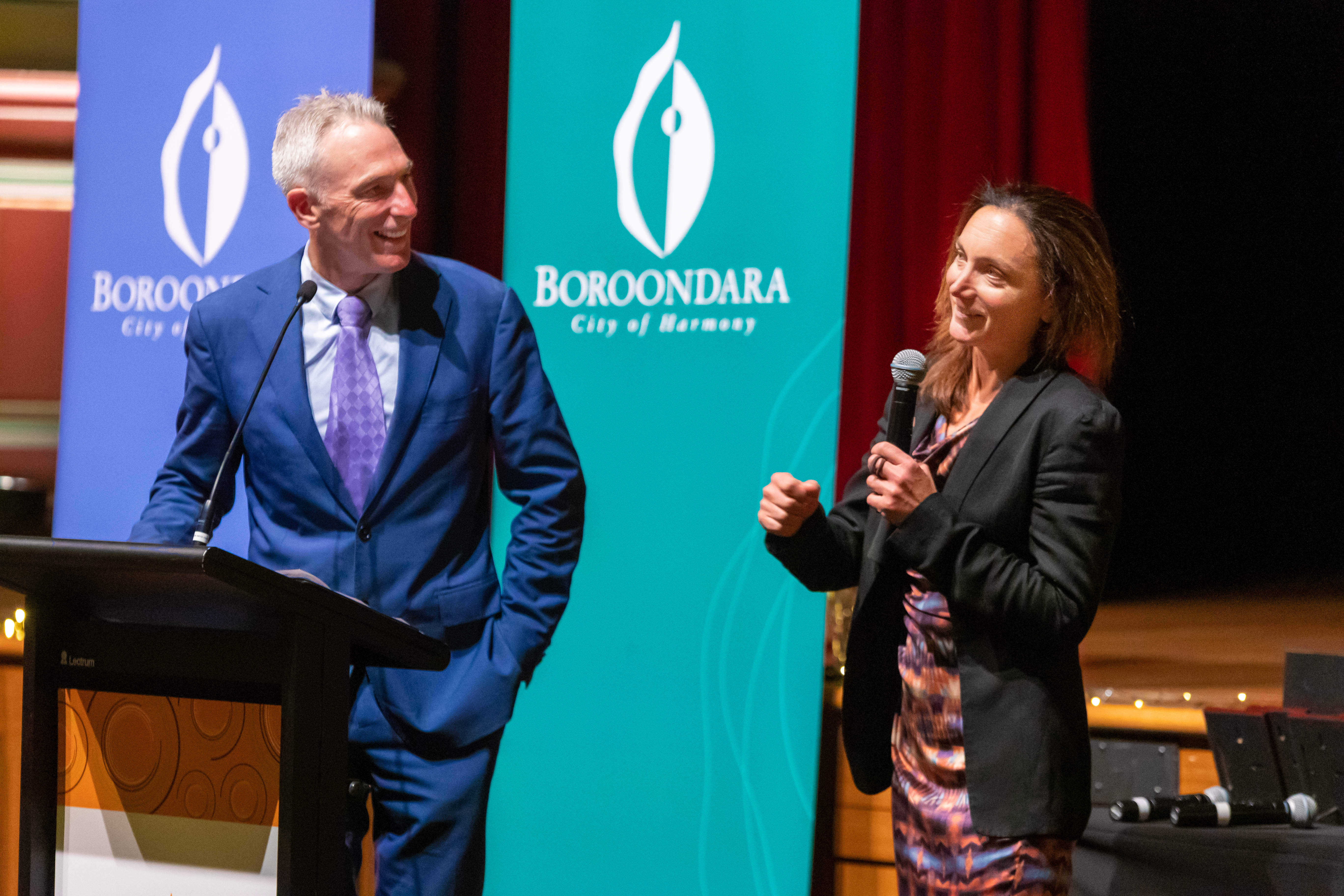 A person standing behind a podium looking at a person speaking with a microphone 