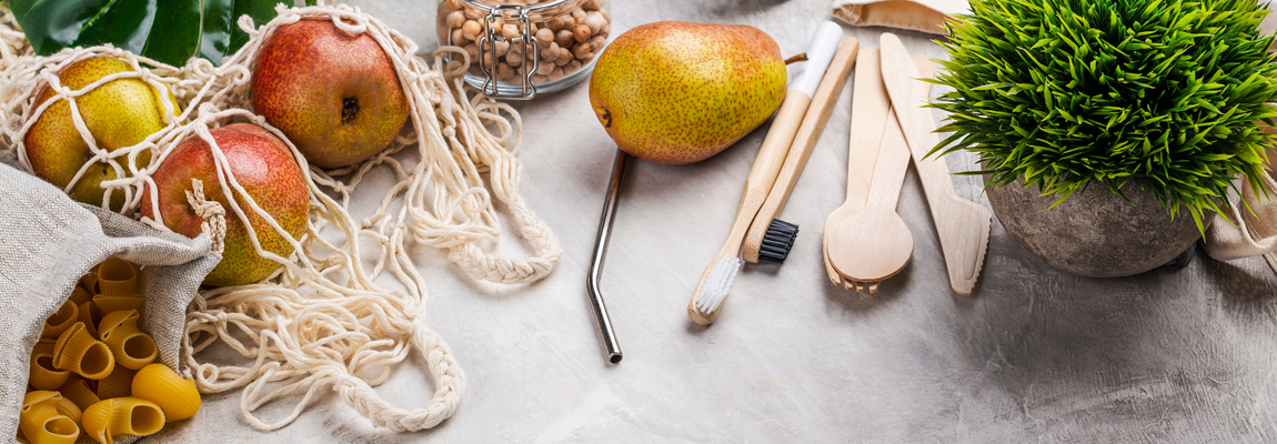 A close-up of a table with pasta, fruit, and nuts all in sustainable containers or bags, and wooden cutlery and toothbrushes with a metal straw and a plant alongside