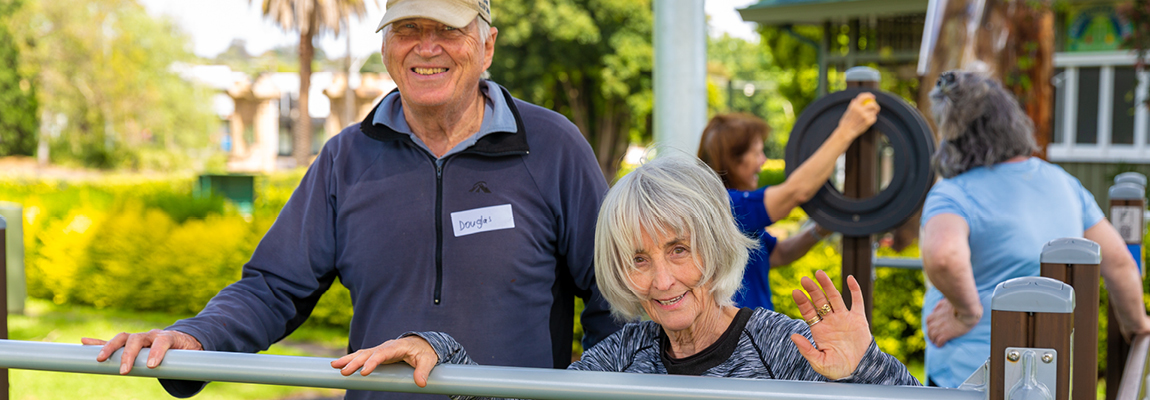 Two older people standing together happily, leaning on some outdoor exercise equipment with people using other equipment behind them