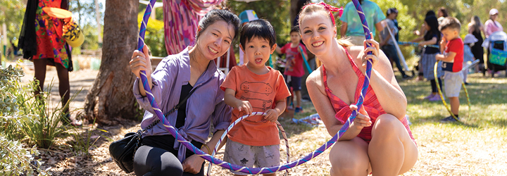 Two adults and a young child looking through a hula hoop on a sunny day in a park with people all around