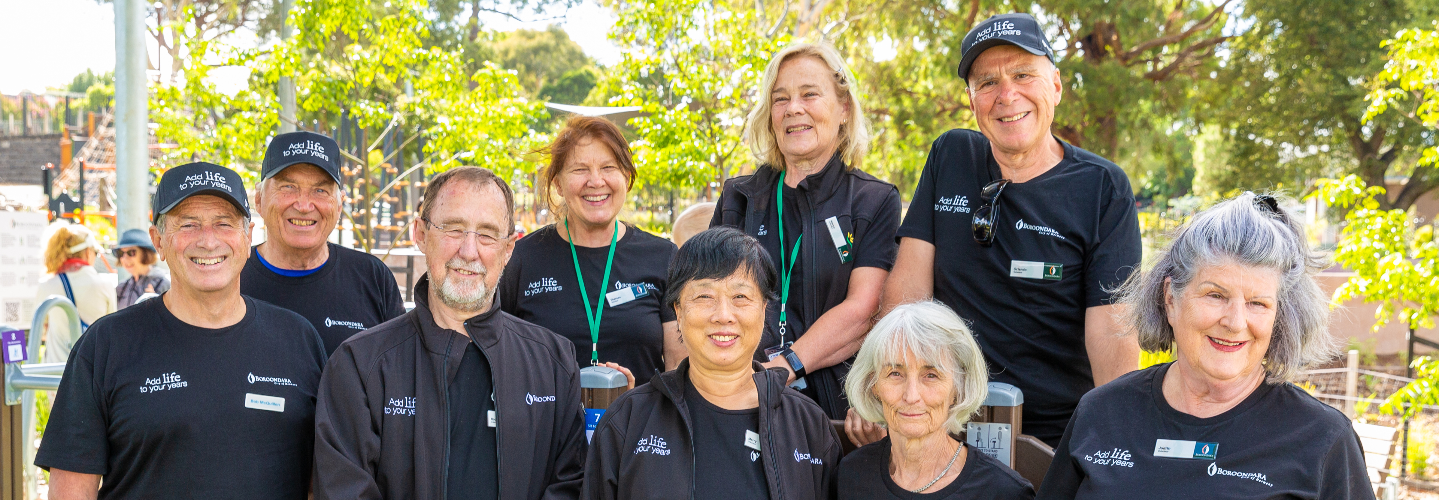 A group of 9 older people who are volunteers at the seniors exercise park program, standing together at the park in their volunteer uniform