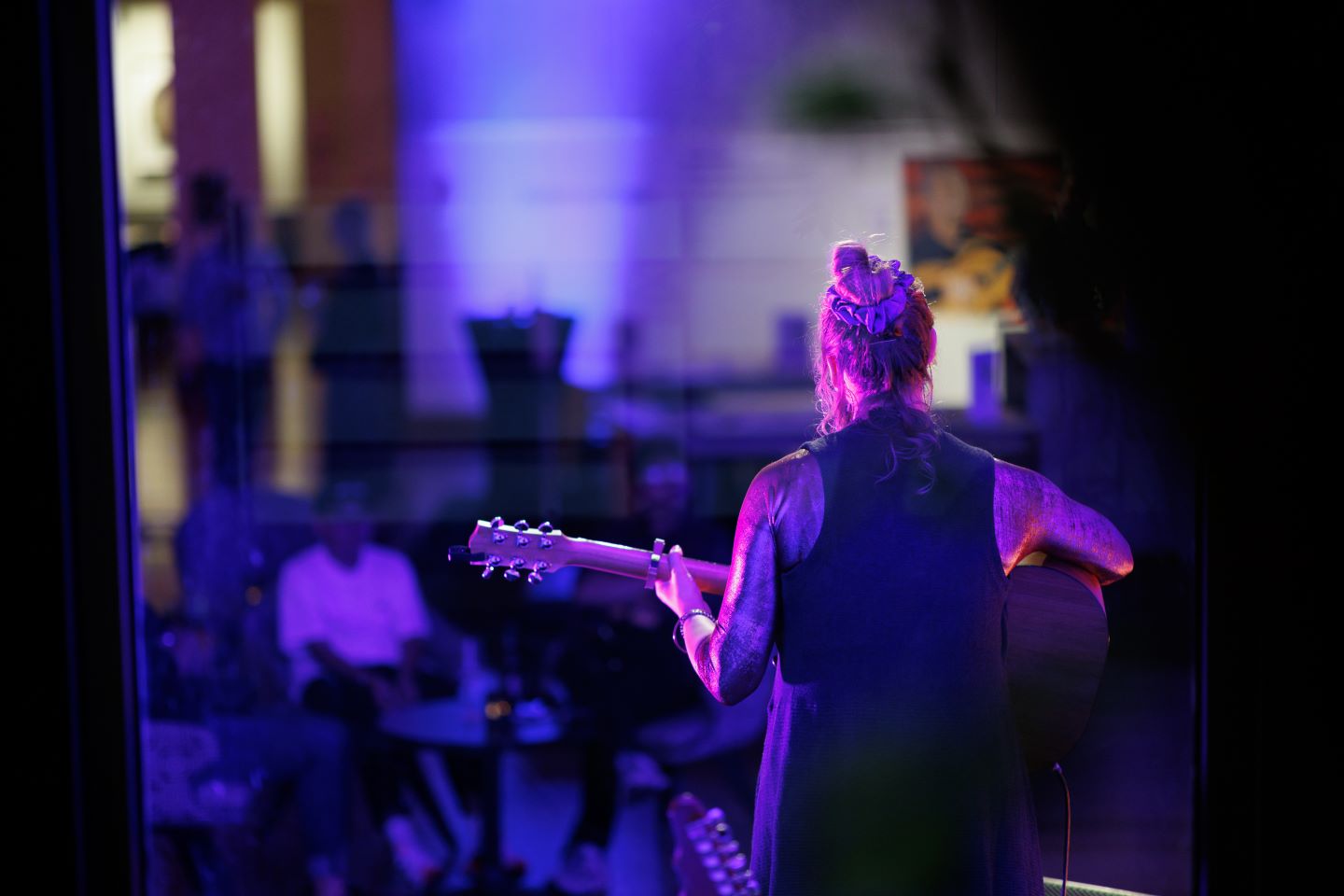 Looking from behind a musician on stage who is lit in purple lighting and holding a guitar, out on the crowd watching the performance