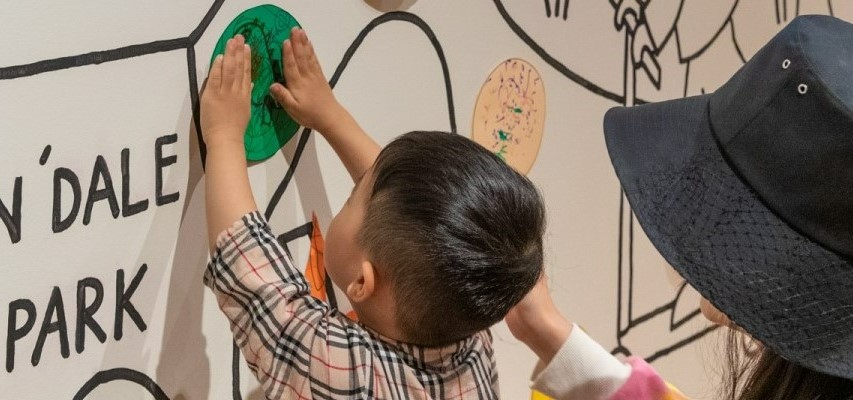 A young child with an adult, adding a piece of craft to a mural on a wall