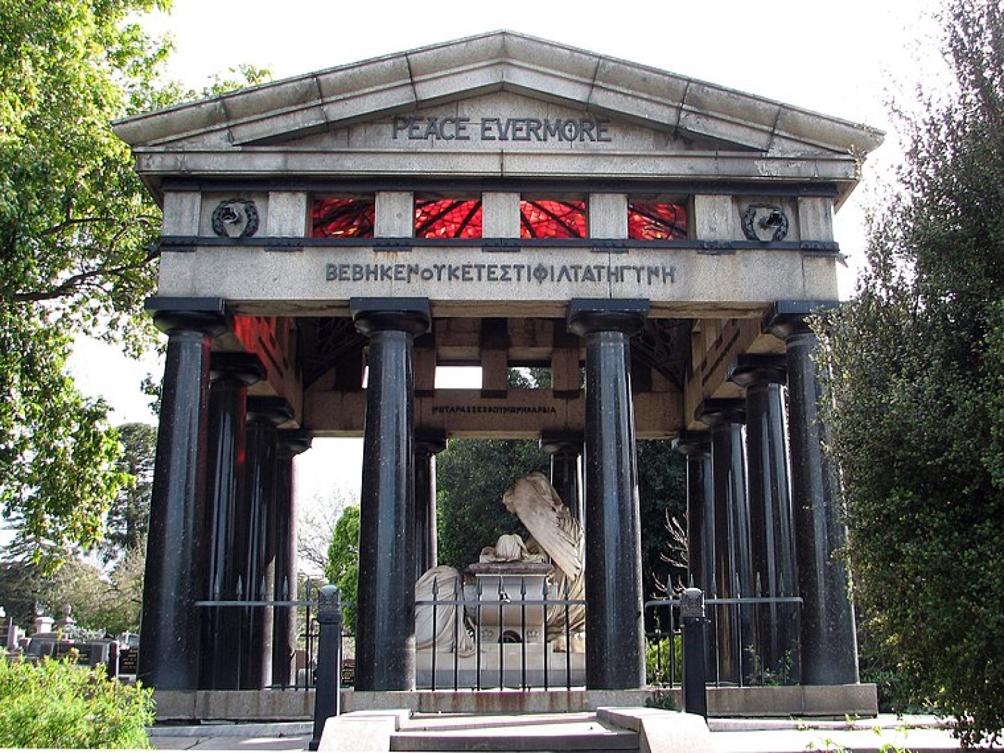 A roman style open air building with coloumns in a cemetary, on the front of the roof it says 'Peace evermore' and there are statues of people inside