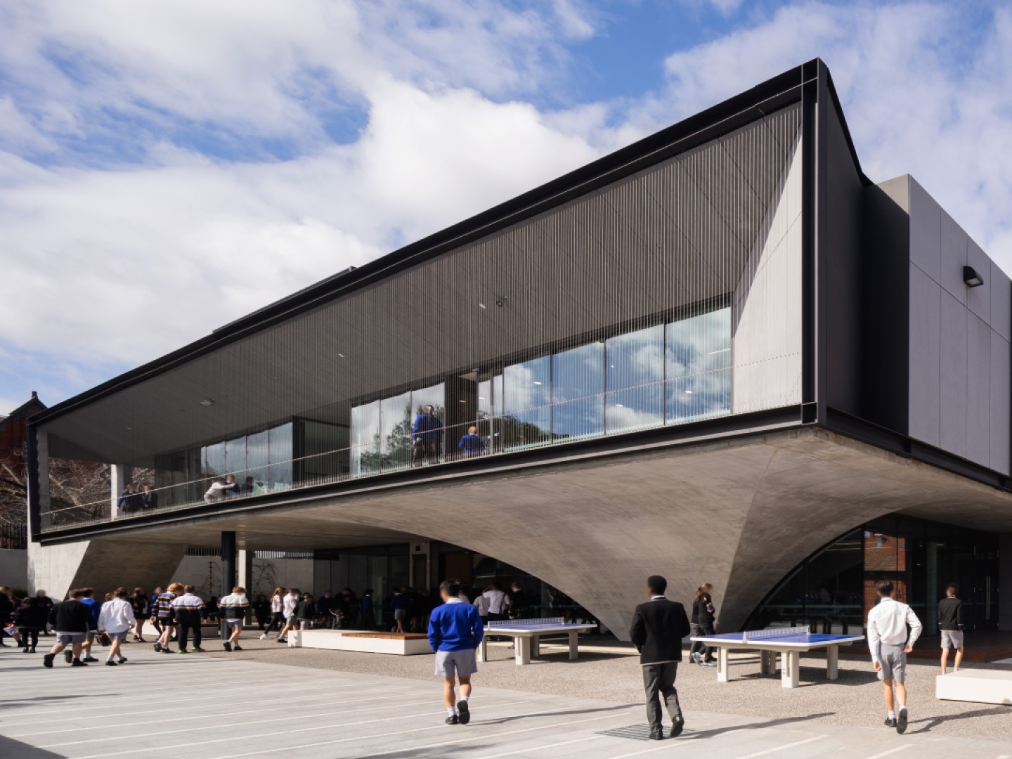 A modern building raised off the ground by wide pillars to make an undercover area, and a second floor with windows looking out over the outdoor area