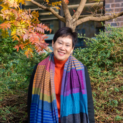 Volunteer Gay Ng in coloured scarf stands in front of tree.