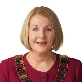 A woman with a blonde bob wears a maroon jacket and ornate gold mayoral necklace