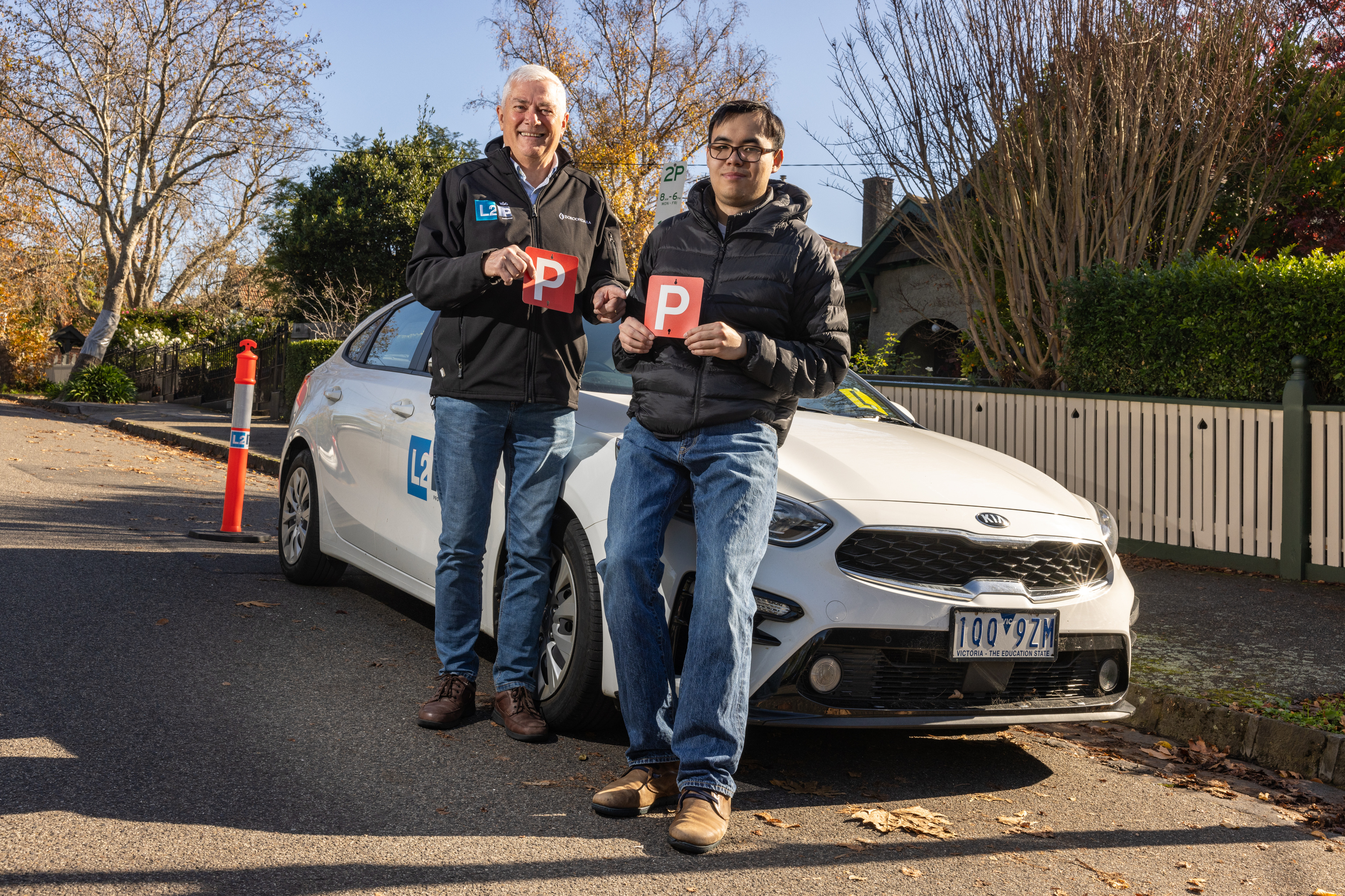 2 men holding P plates stand next to a white car