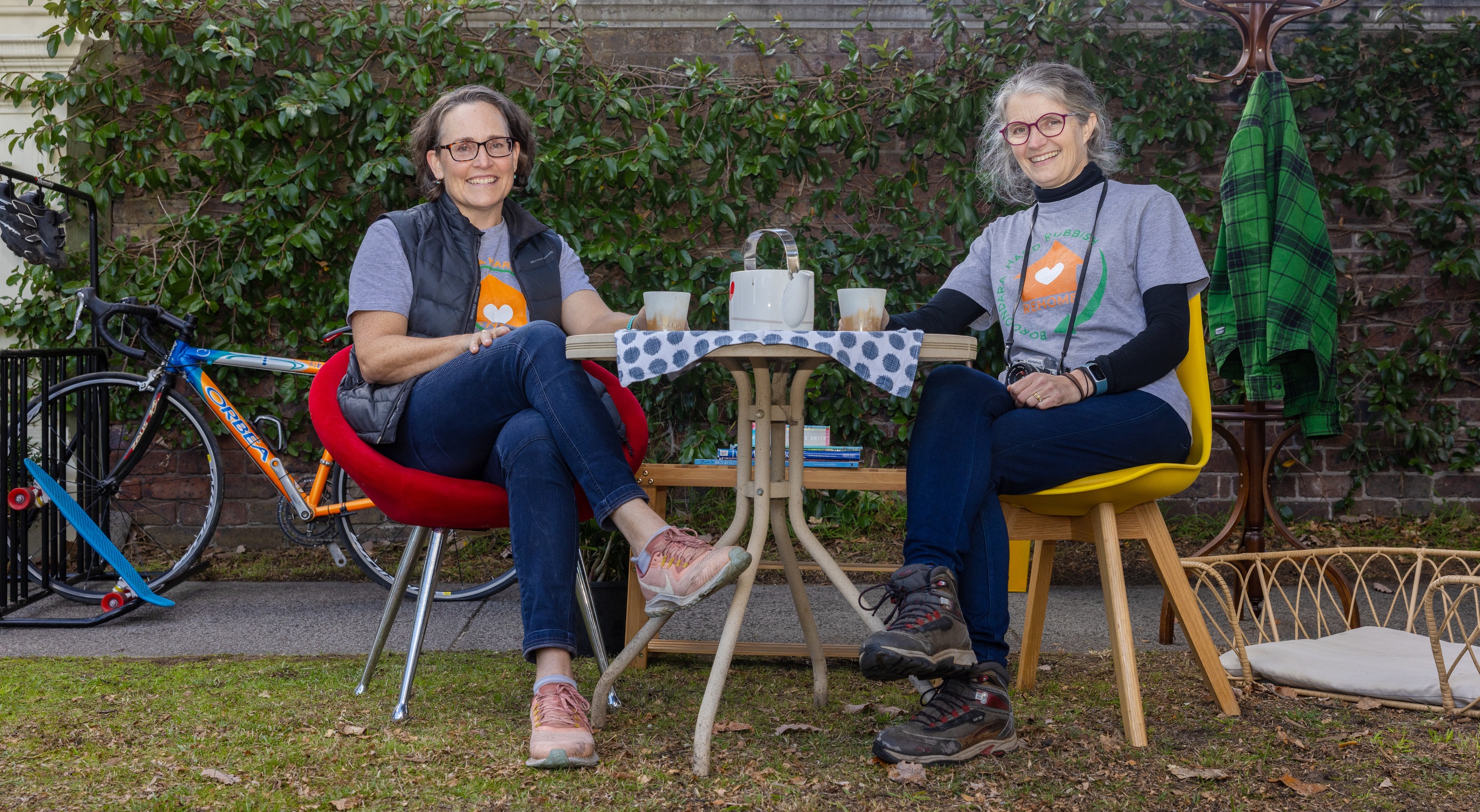 2 smiling women sit at an outdoor table with bikes parked behind them