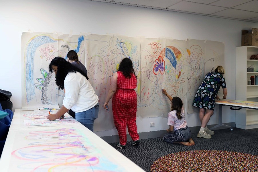 Several young people in a room. They are drawing colourful artwork on butchers paper on tables and walls in the room.