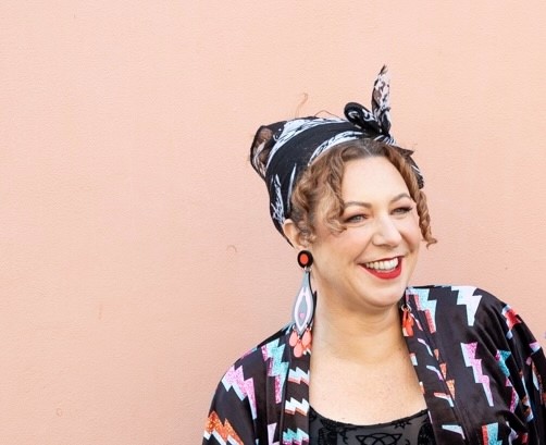 woman with curly brown hair hanging out from under a headscarf. She is wearing big dangly earrings and a black top with coloured lightning bolts on it