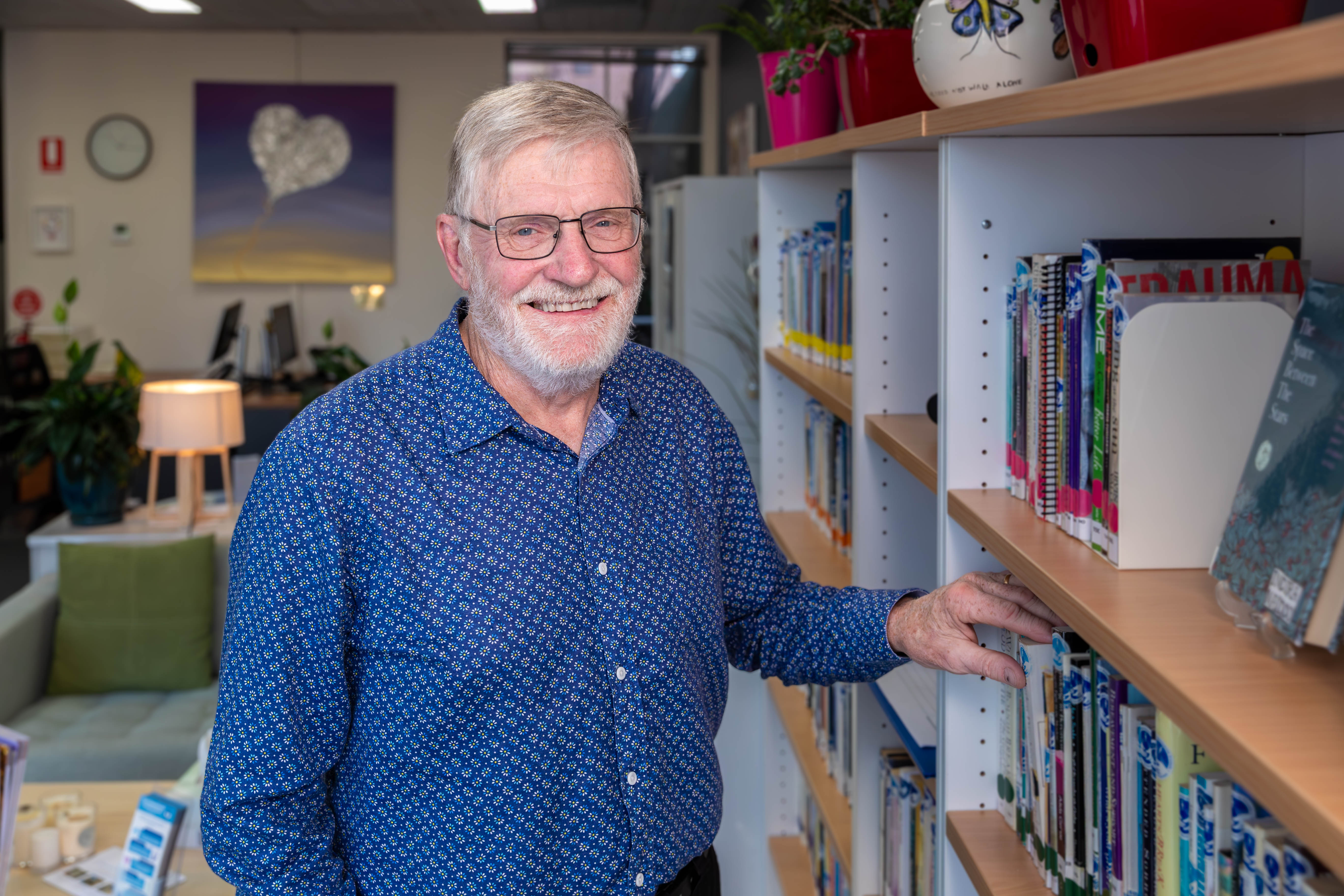 grey haired man wearing a blue shirt, standing at a bookcase