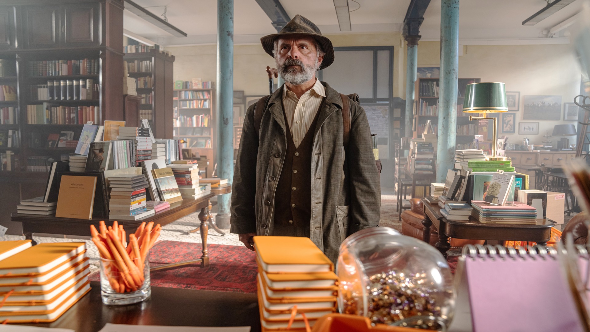 A man in a well-worn hat and suit stands in a bookstore