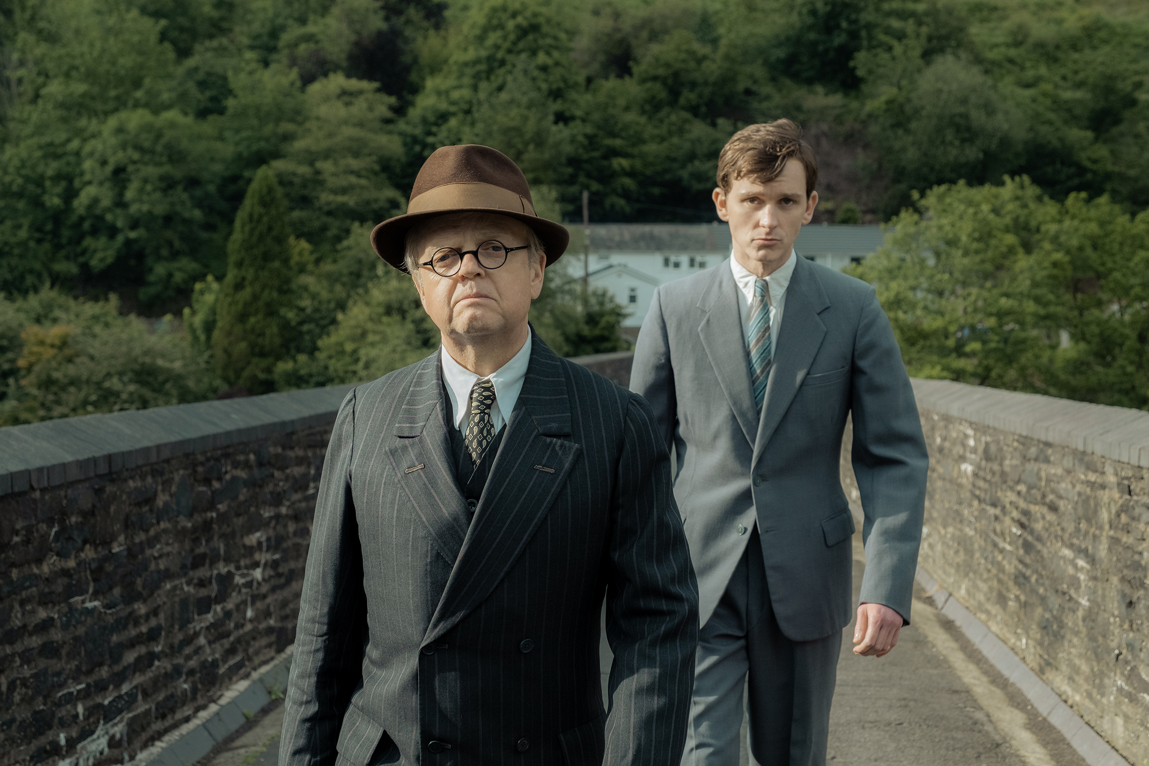 One man in hat, suit and tie is followed by another in a suit on a stone bridge
