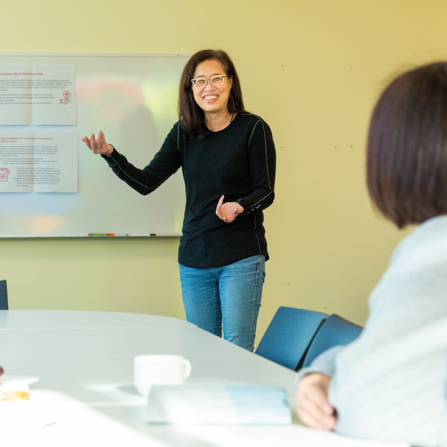 A woman in a black top pointing at a whiteboard at the front of a room