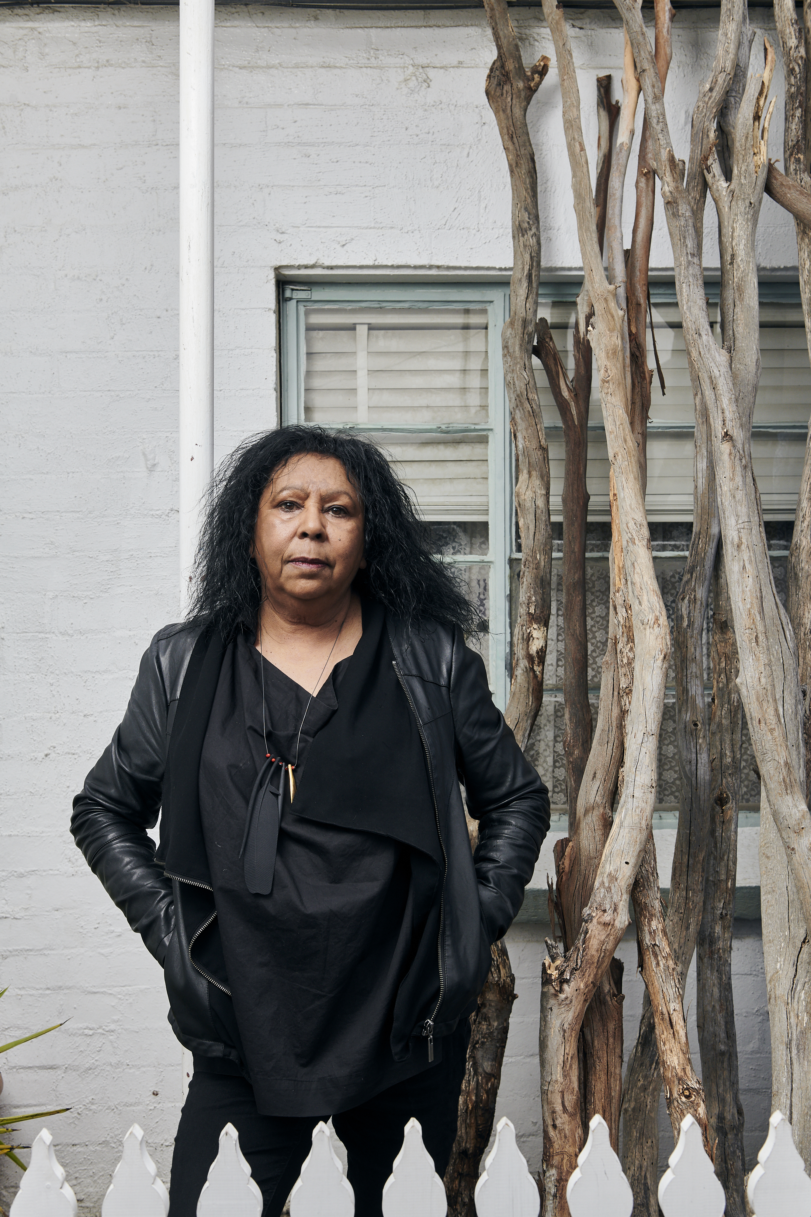 A woman wearing black clothing stands in front of a house and next to a tree.