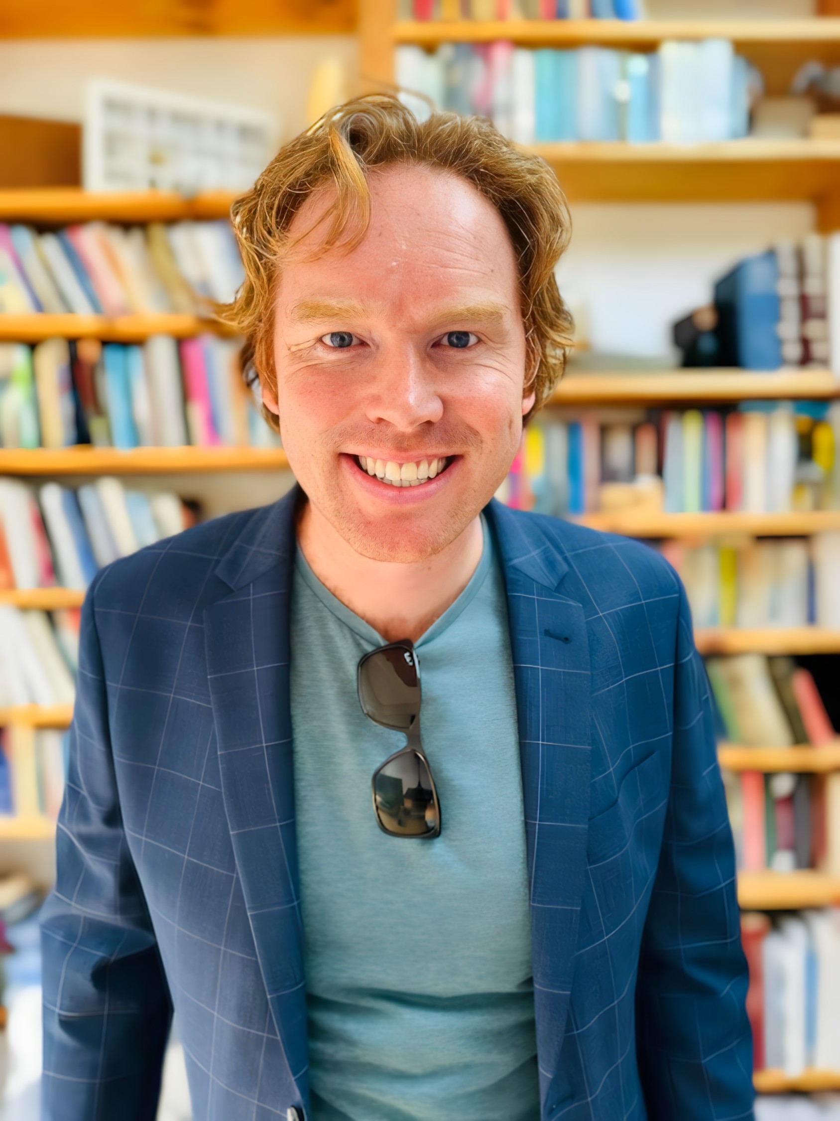 Man with red hair wears a green tshirt with sunglasses hanging from the collar, and a blue jacket smiles. In the background are shelves full of books.