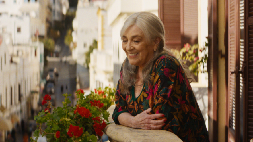 An older woman leans on the railing of a balcony, looking down to the street below. She is smiling.