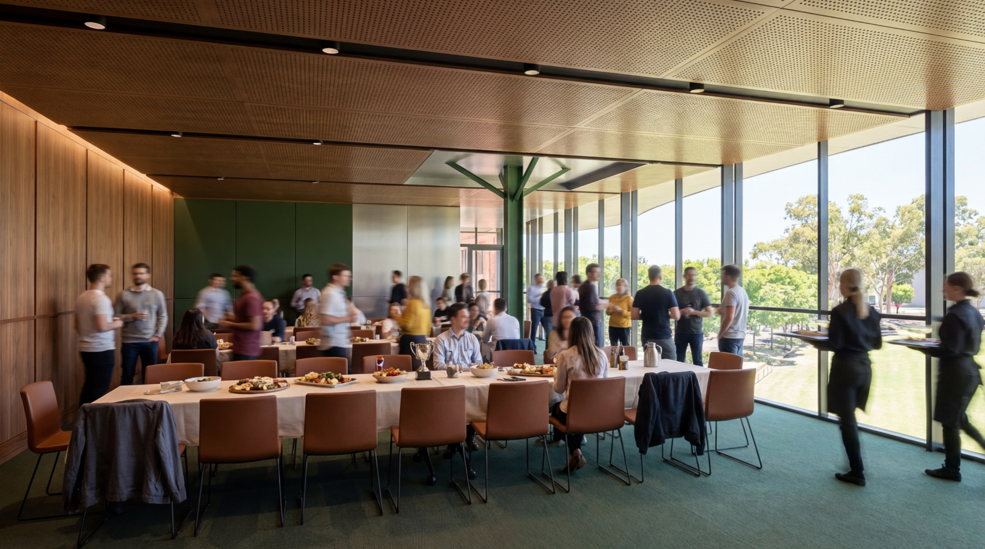 A large multipurpose space with carpet and wood panneling looks over the ground through large windows.The space is currently filled with 2 large tables and people milling about while food is being served.