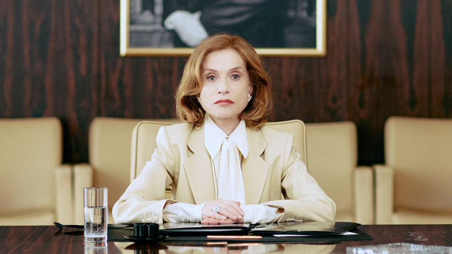 a woman with shoulder length red hair wearing a cream suit jacket sits at a boardroom table with folders in front of her.