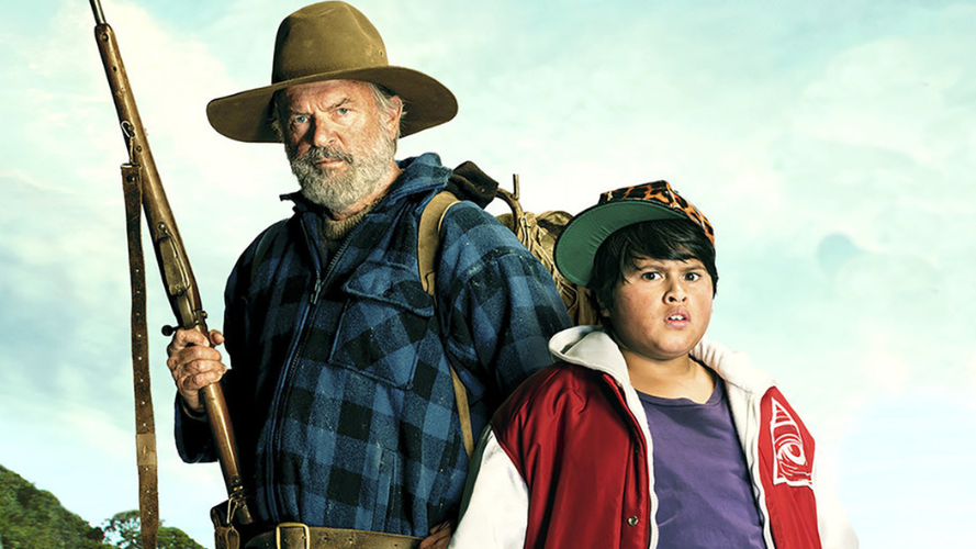 An older man wearing a flannellette shirt and wide brimmed hat holds a gun. next to him stands a young teenager in a red bomber jacket arms out in a 'what' gesture.