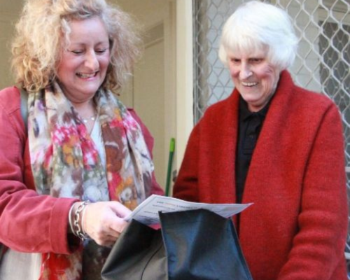 A volunteer delivering a bag of books to a person at their front door as part of the Home Library Service. Both are smiling as the volunteer checks a delivery note.
