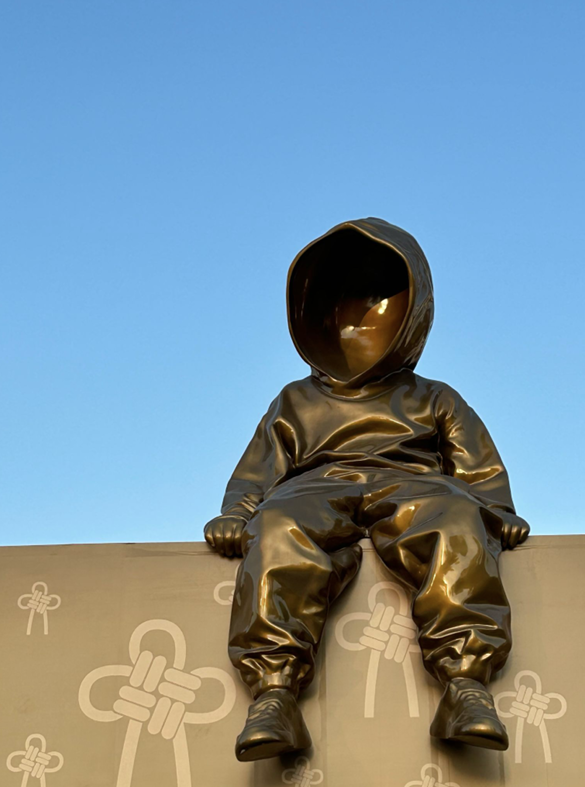 Photograph of brass statue of a person wearing a hoodie sitting on a wall