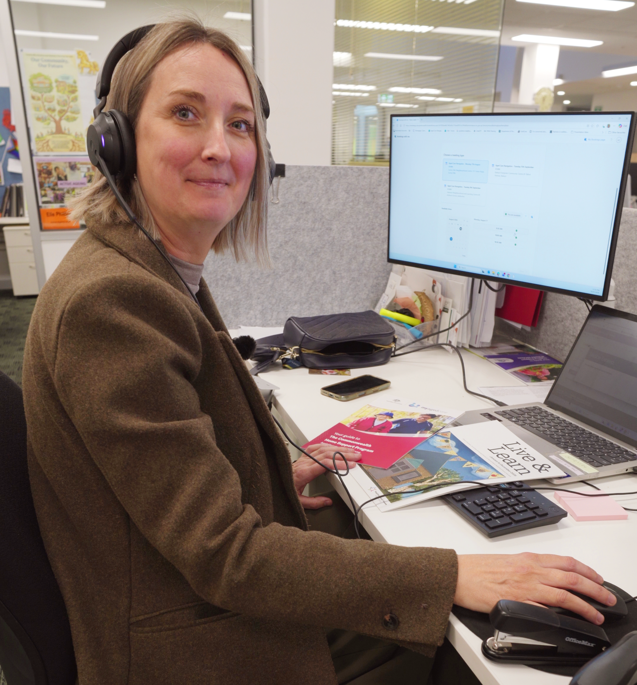 Woman in a brown coat sits in front of a computer at a desk.