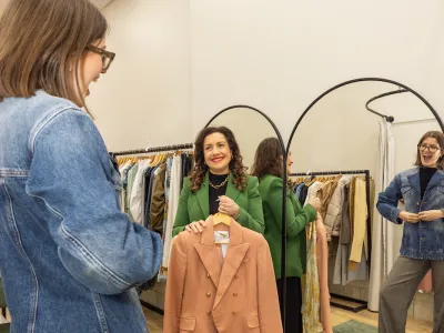 2 women in a clothing store, one is the sales assistant holding up a jacket while the other is a customer trying on a jacket.