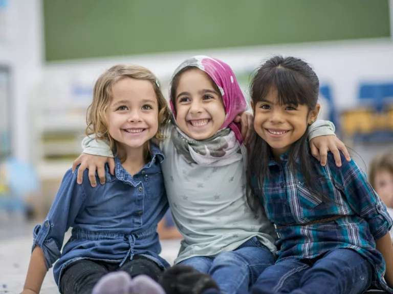 Three young girls smile together. The middle girl has her arms over the other two girls.