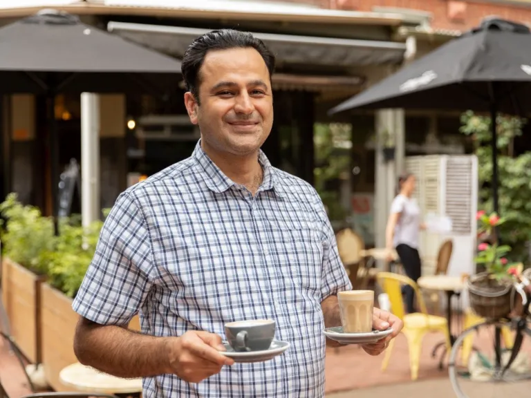 A local business owner stands outside a cafe holding two coffees
