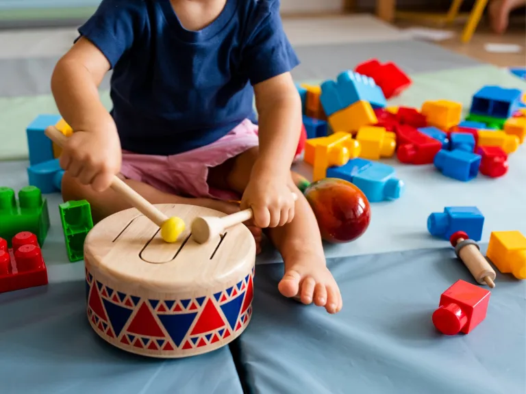 Toddler playing with a wooden toy drum. There are plastic building blocks scattered across the floor around them.